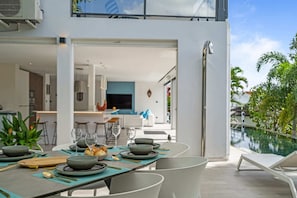 A modern dining area with a pool view, featuring a table set for a meal, a kitchen in the background, and a clear sky.