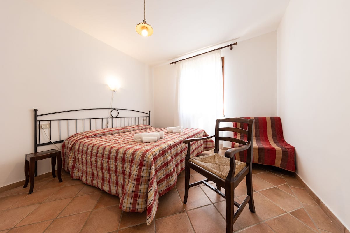 A bedroom with a bed, a chair, a small table, and a window with curtains.