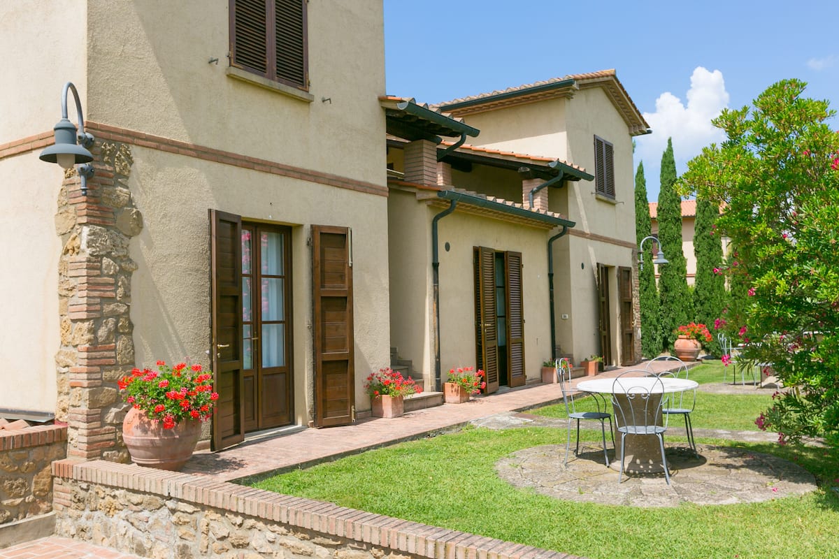 A two-story building with a stone facade, multiple windows with shutters, and a small patio area with a table and chairs.