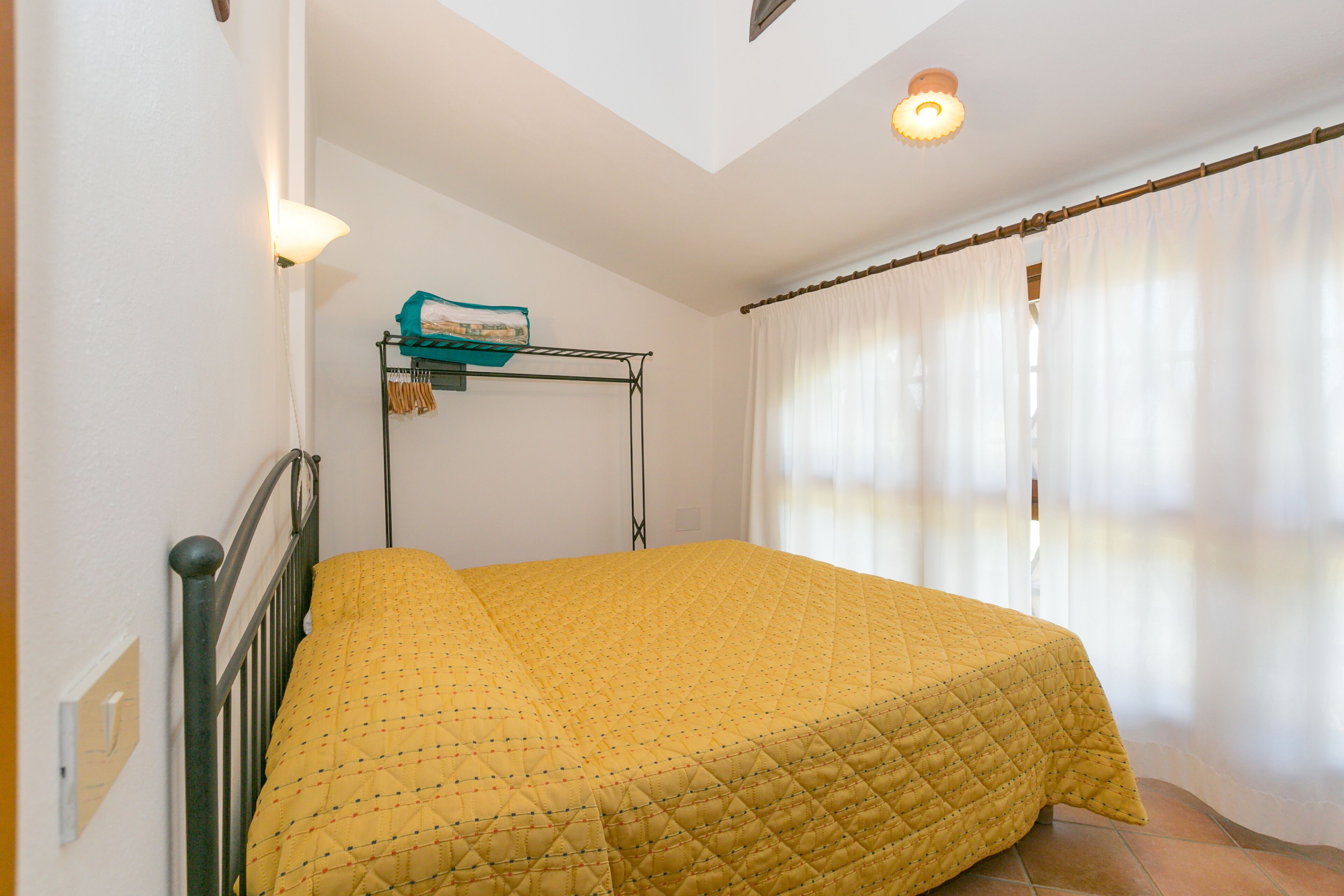 A bedroom with a bed, a wall-mounted shelf with items, and a window with curtains.