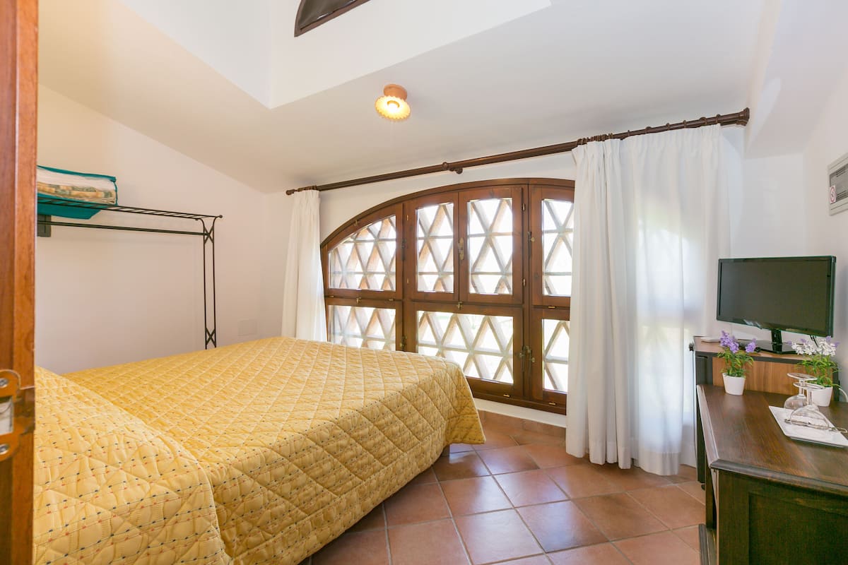 A bedroom with a bed, a TV, a window with curtains, and a desk with a plant.