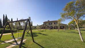 Un área verde con un conjunto de columpios, un sendero y una casa grande al fondo.