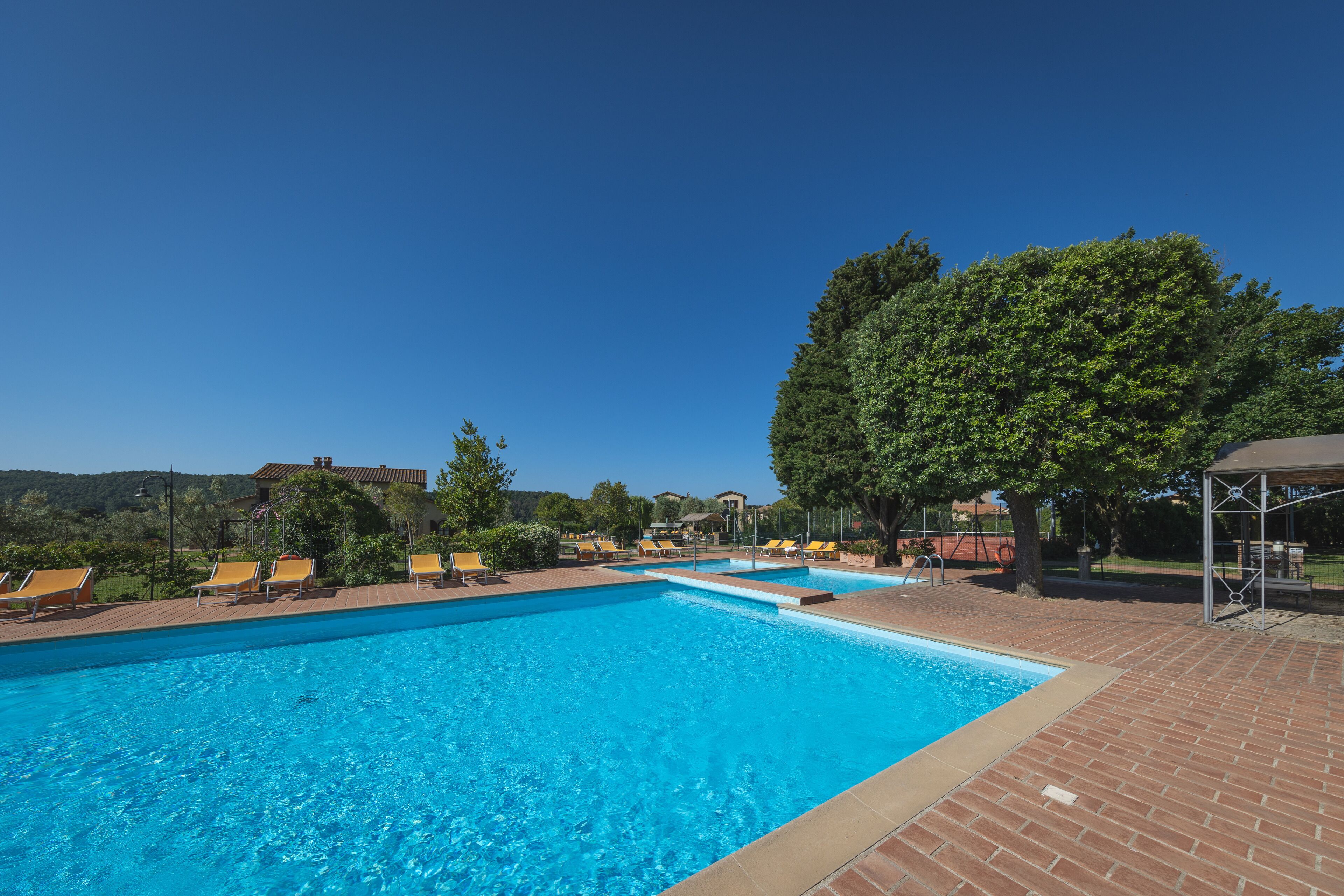 A rectangular swimming pool with clear blue water, surrounded by a brick patio and yellow lounge chairs.