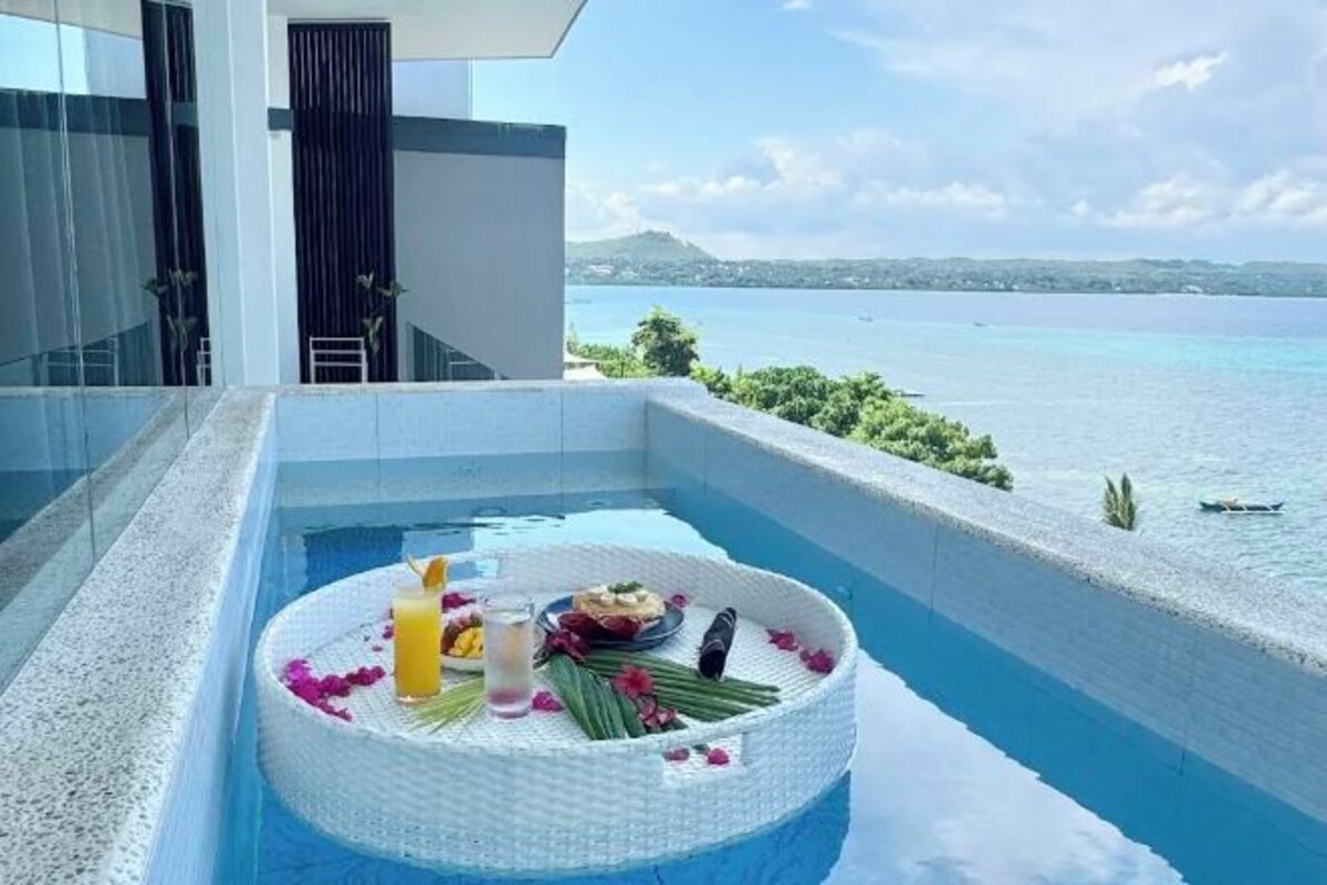 A modern infinity pool with a floating platform containing a cake, candles, and flowers, overlooking a clear blue sea and distant land.