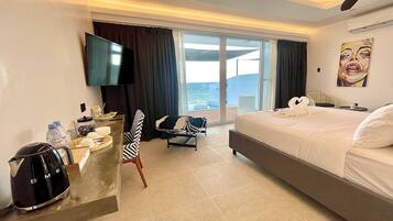 A hotel room with a bed, a desk with a kettle, a chair, a TV, and a view of the sea.