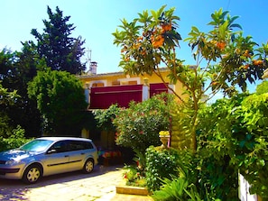A residential area with a parked car, a building with a red shutter, and lush greenery.