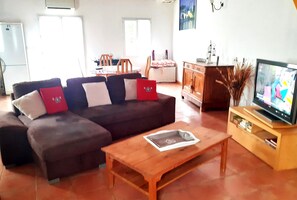 Una sala de estar con un sofá modular, una mesa de centro de madera, un televisor sobre un mueble de madera y un comedor al fondo.