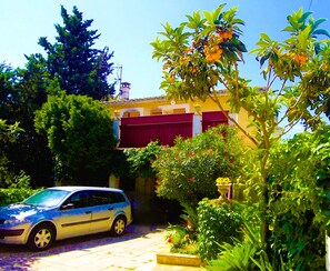 Zona residencial con un automóvil estacionado, un edificio con persiana roja y abundante vegetación.
