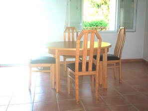 Un coin repas comprenant une table ronde en bois et quatre chaises, un sol carrelé et une fenêtre donnant sur un paysage verdoyant.