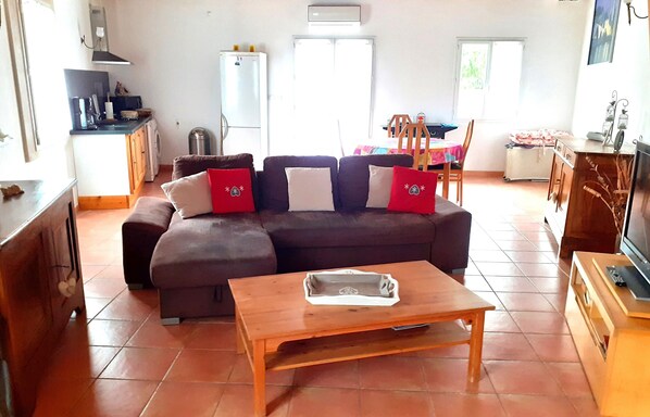 Una sala de estar con un sofá marrón, una mesa de centro de madera, una zona de cocina con un refrigerador y una mesa de comedor con sillas.
