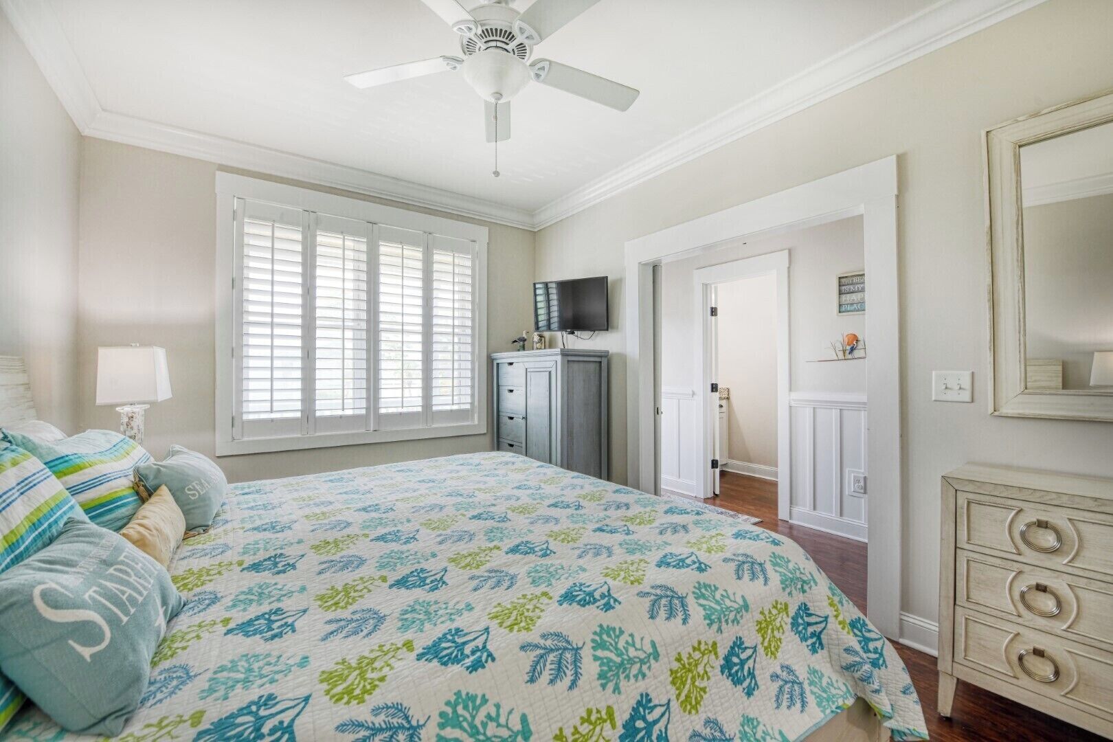 A bedroom with a bed, a ceiling fan, a dresser, a television, and a door leading to another room.