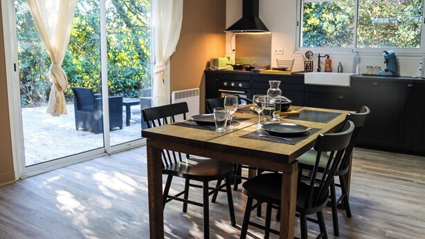 A dining area with a table set for four, a kitchen in the background, and a view of a snowy garden through glass doors. - Duplex Le Philandra - 2-4 pers - jacuzzi  (Monteils)