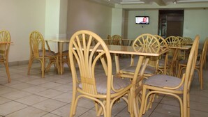 A dining area with a long table and wicker chairs, a television on the wall, and a tiled floor. - Roof Galaxy Hotel (Machakos)