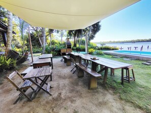 An outdoor patio with wooden tables and benches, a barbecue grill, and a view of a swimming pool and lake.