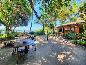 A beachside patio with a wooden table and chairs, surrounded by lush greenery and a view of the sea.