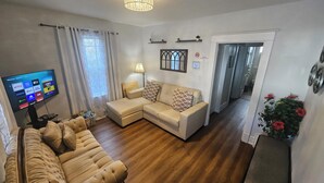 Een woonkamer met een beige bank, een aan de muur bevestigde televisie, een lamp en een raam met gordijnen.