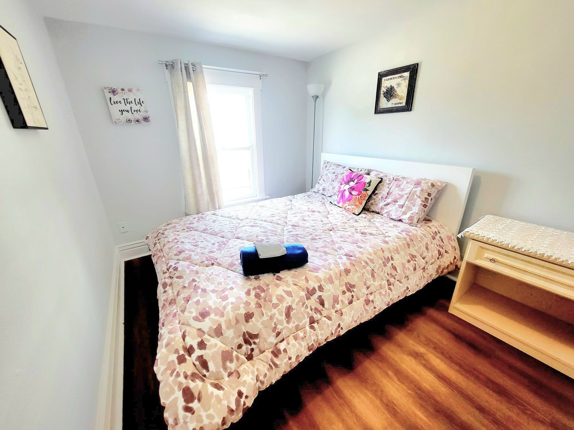 Un dormitorio con una cama, una mesita de noche, una ventana con cortinas, un cuadro en la pared y un cartel en la pared.