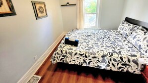 Een slaapkamer met een bed, een raam met gordijnen, een plank met een plant en twee ingelijste foto's aan de muur.