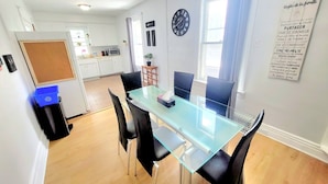 Een eethoek met een glazen tafel en zwarte stoelen, een klok aan de muur en een poster met huisregels.