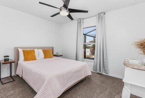 A bedroom with a bed, a ceiling fan, a window with curtains, a bedside table with a plant, and a small desk with a basket. - SunnyBeach Luxury Vacation House*Pool*Spa*Central Location (Fort Myers)