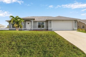 A modern single-story house with a garage, a well-maintained lawn, and a palm tree. - SunnyBeach Luxury Vacation House*Pool*Spa*Central Location (Fort Myers)
