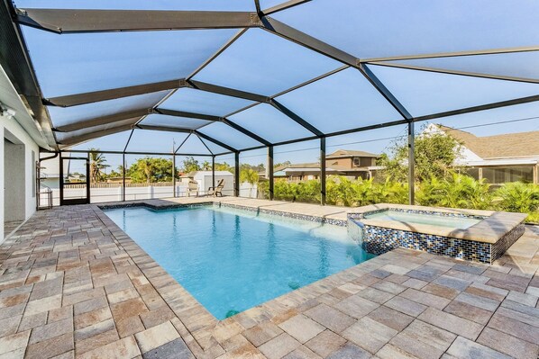 A covered swimming pool with a tiled edge and a hot tub, surrounded by a brick patio and a metal roof structure. - SunnyBeach Luxury Vacation House*Pool*Spa*Central Location (Fort Myers)