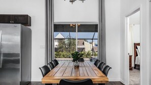A dining area with a wooden table and chairs, a modern stainless steel refrigerator, and a view through large windows.