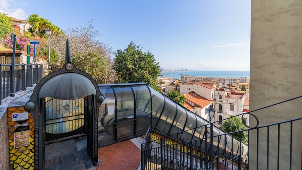 A staircase with a glass enclosure leading to a higher floor, overlooking a coastal cityscape. - Panacea (Salerno)