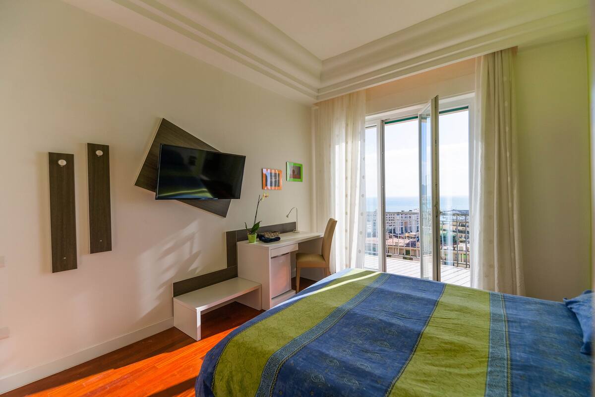 A hotel room with a bed, a desk, a TV, and a balcony with a city view.