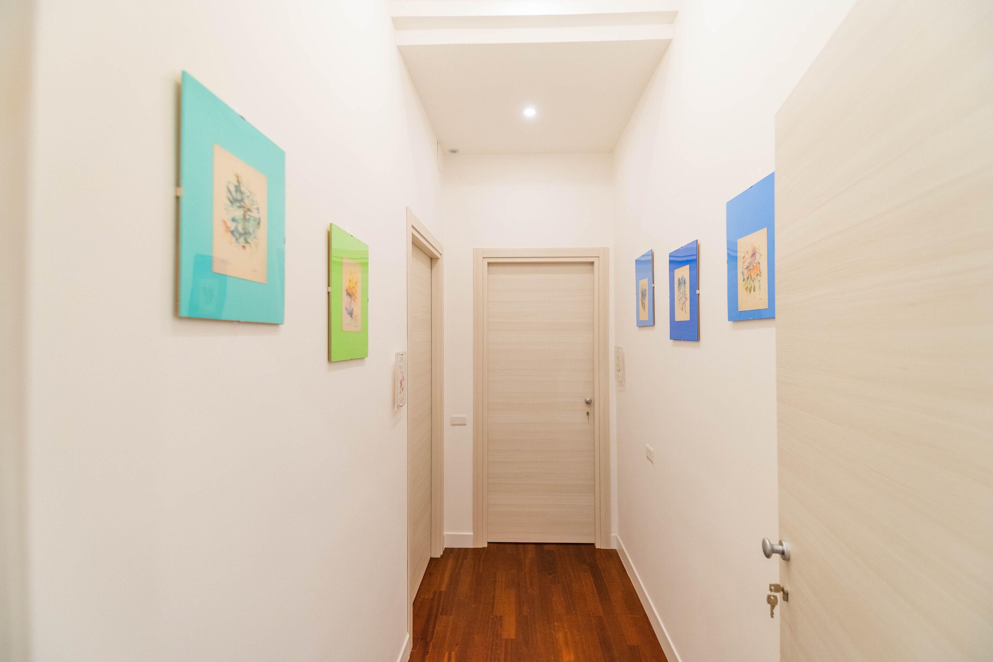 A narrow hallway with wooden flooring, white walls, and several framed artworks on the walls.