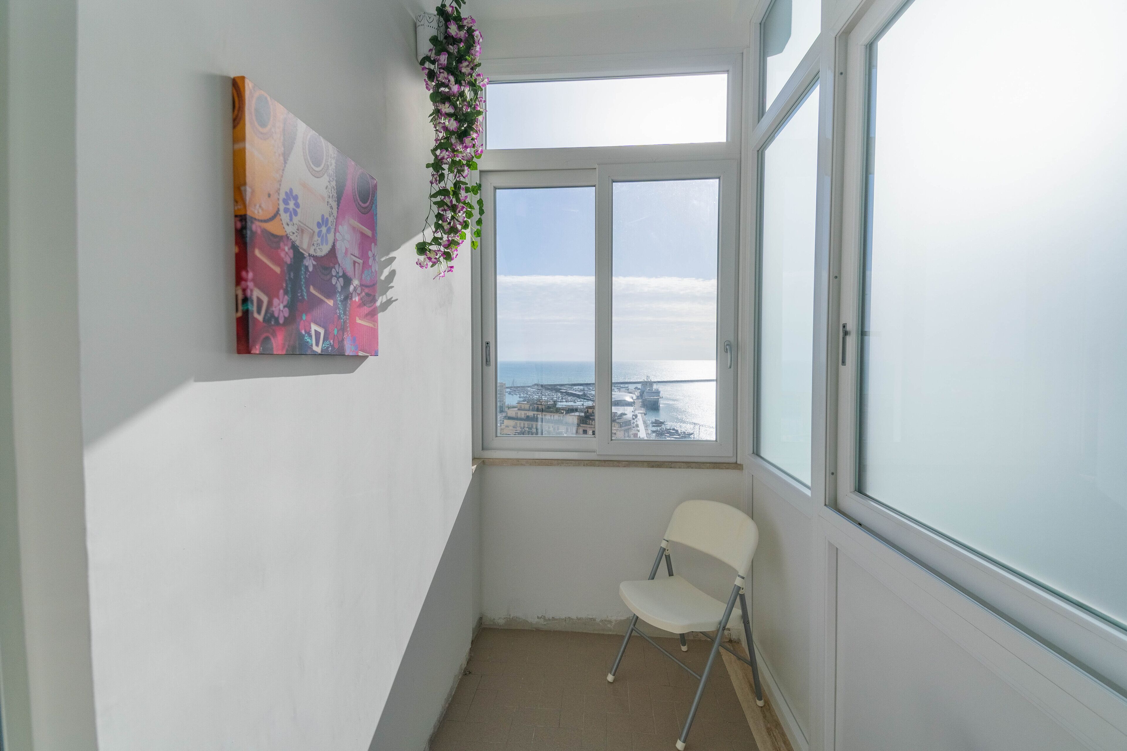 A narrow room with a large window, a white chair, and a colorful painting on the wall.
