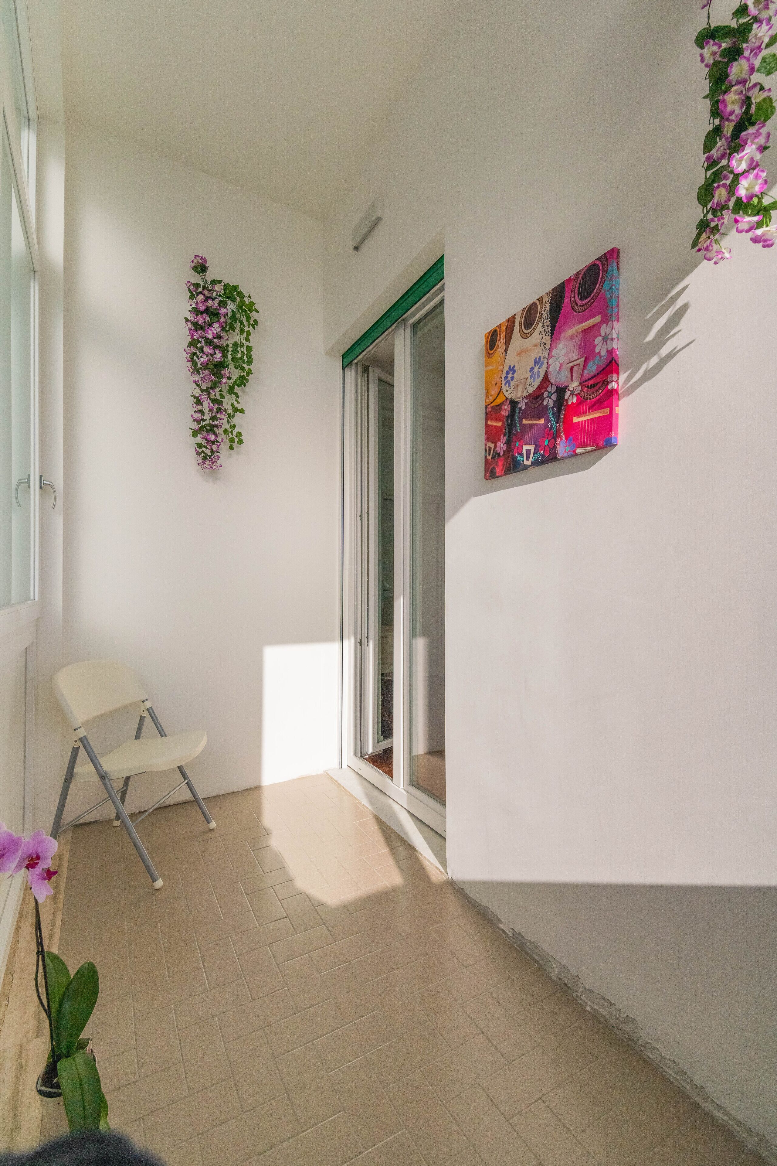 A balcony with a white chair, a hanging plant, and a colorful painting on the wall.