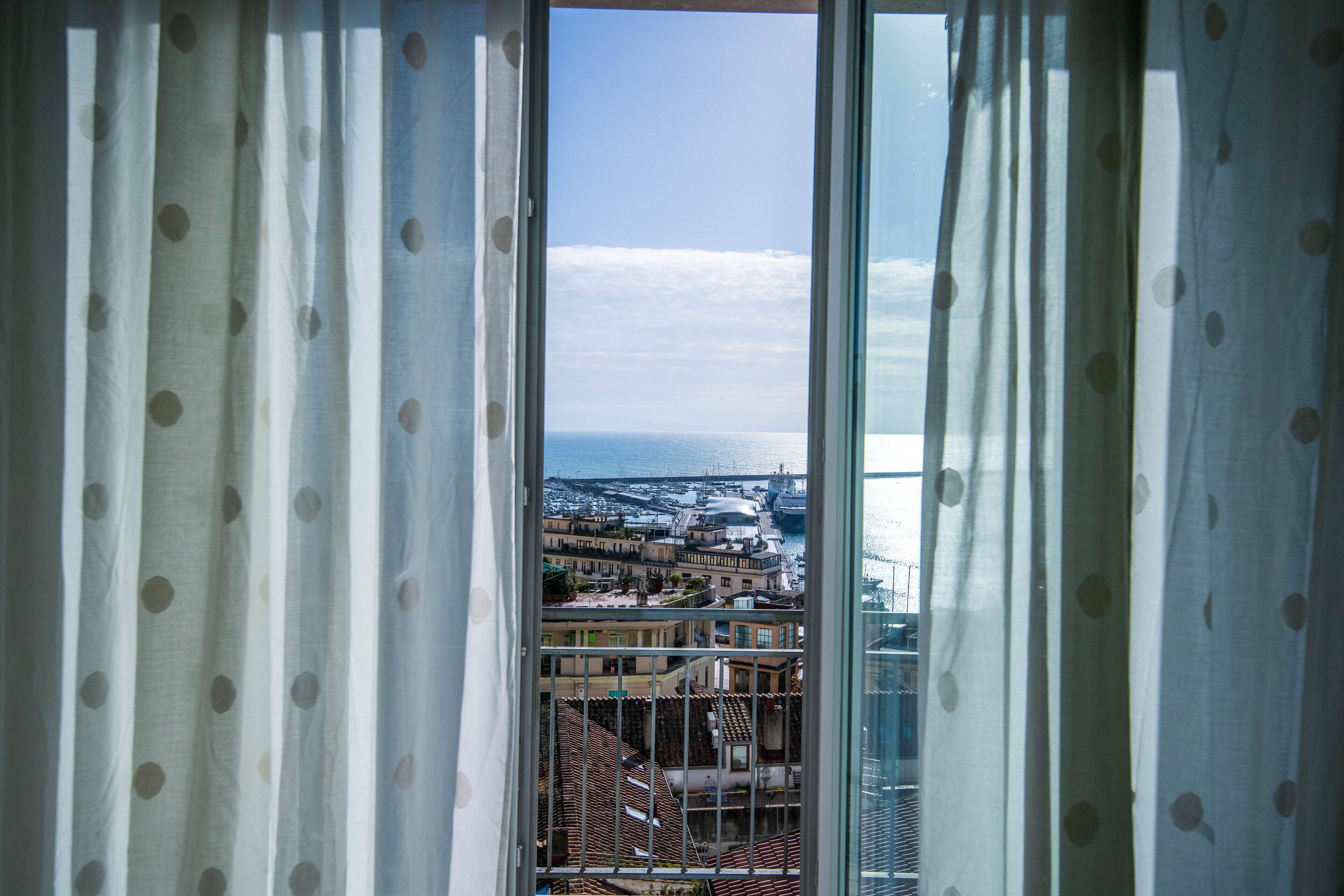 Vista a través de una ventana con cortinas de lunares, mostrando un paisaje urbano costero con edificios y un puerto.