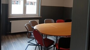 Een vergaderruimte met een ronde tafel, zes stoelen, een groot raam en een radiator.
