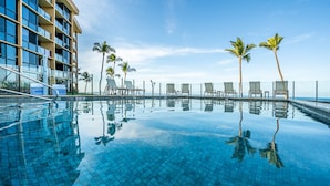 Une magnifique piscine à débordement d’un bleu profond, bordée de chaises longues et de palmiers, qui reflète un bâtiment moderne doté de balcons.