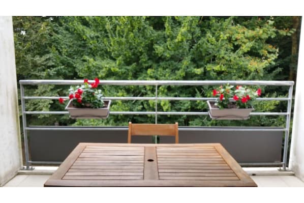 A balcony with a wooden table and two potted plants with red flowers.