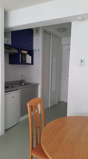 A compact kitchen with a sink, stove, and cabinets, a dining table with a chair, and a door leading to another room.