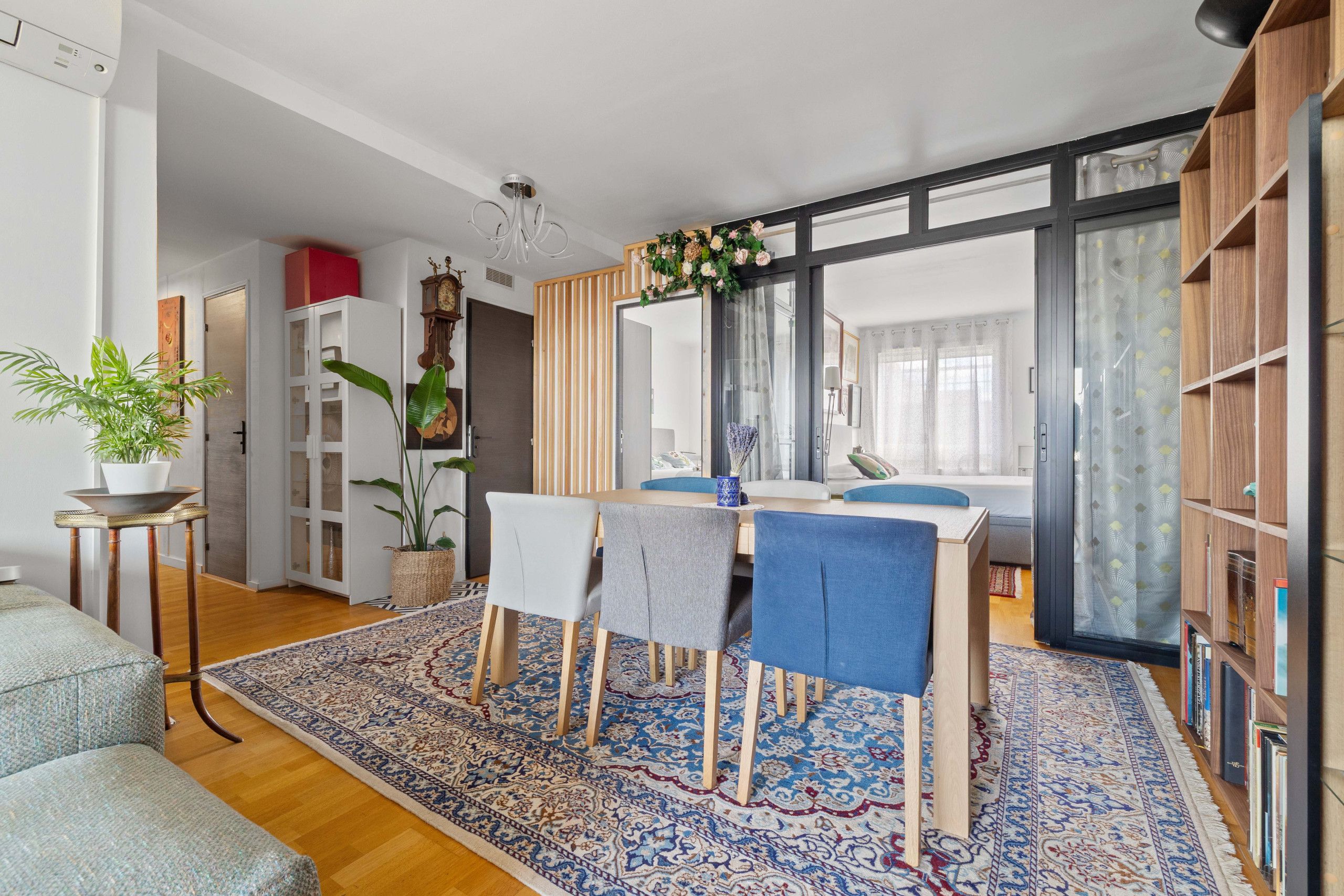 A modern living room with a dining area, wooden flooring, a large window, and a bookshelf.