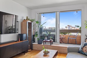 A modern living room with a flat-screen TV, wooden furniture, and a large window offering a city view. - Attica, 3BR/2BA, 6 people (París)