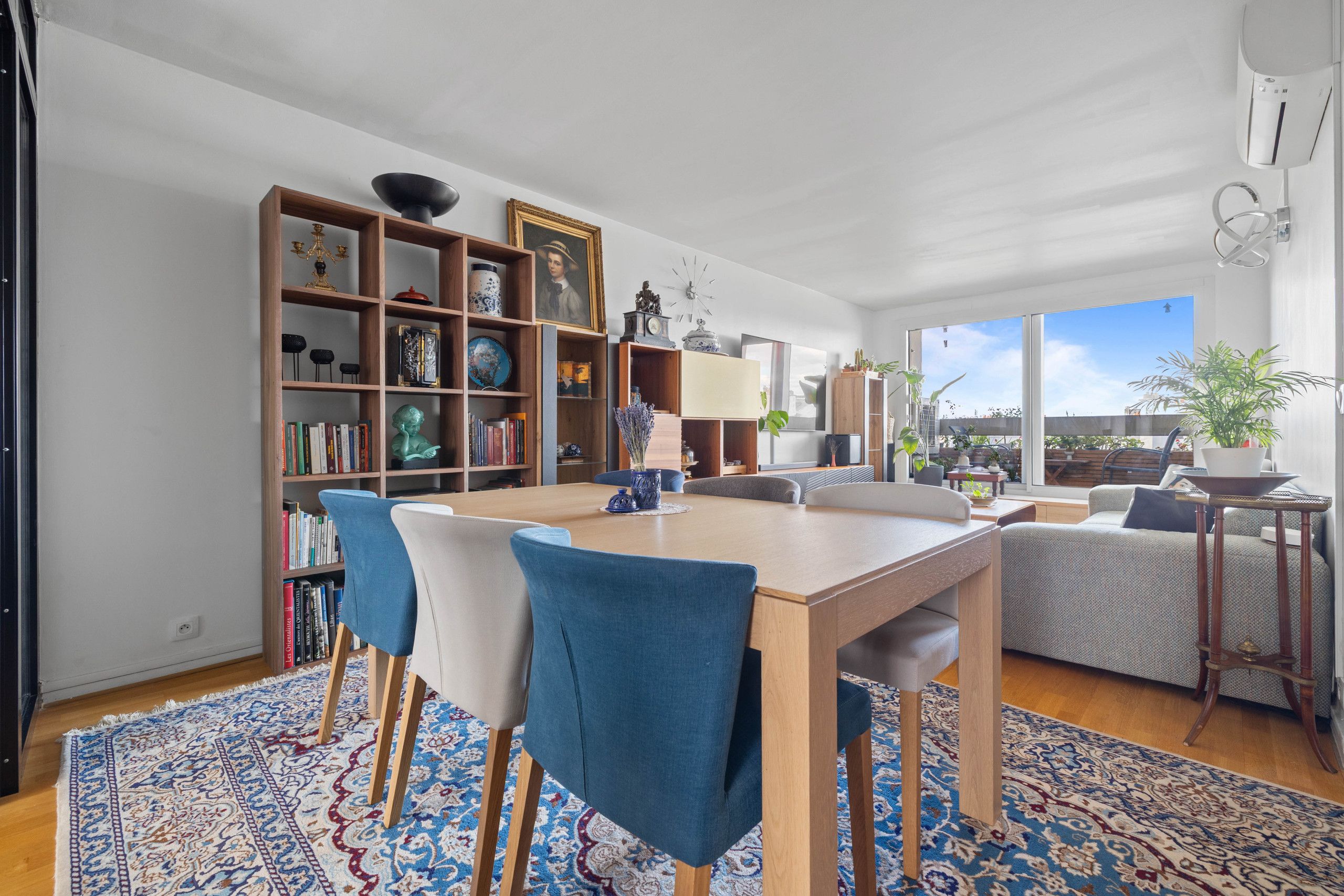 Een moderne woonkamer met een eethoek, met een houten tafel, blauwe stoelen, een boekenkast en een groot raam met uitzicht.