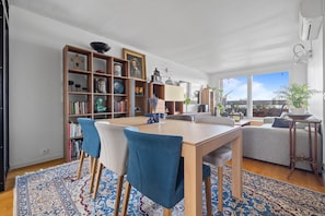 Un salon moderne avec un coin repas, comprenant une table en bois, des chaises bleues, une bibliothèque et une grande fenêtre donnant sur un paysage.