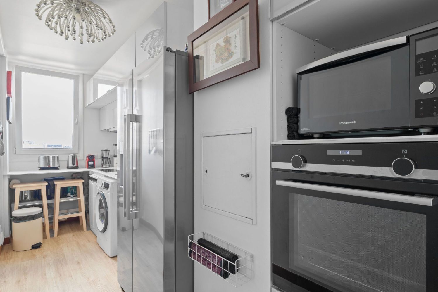 A compact kitchen with a microwave, oven, and refrigerator. There is a small table with a kettle and a coffee maker. A washing machine is visible in the background.