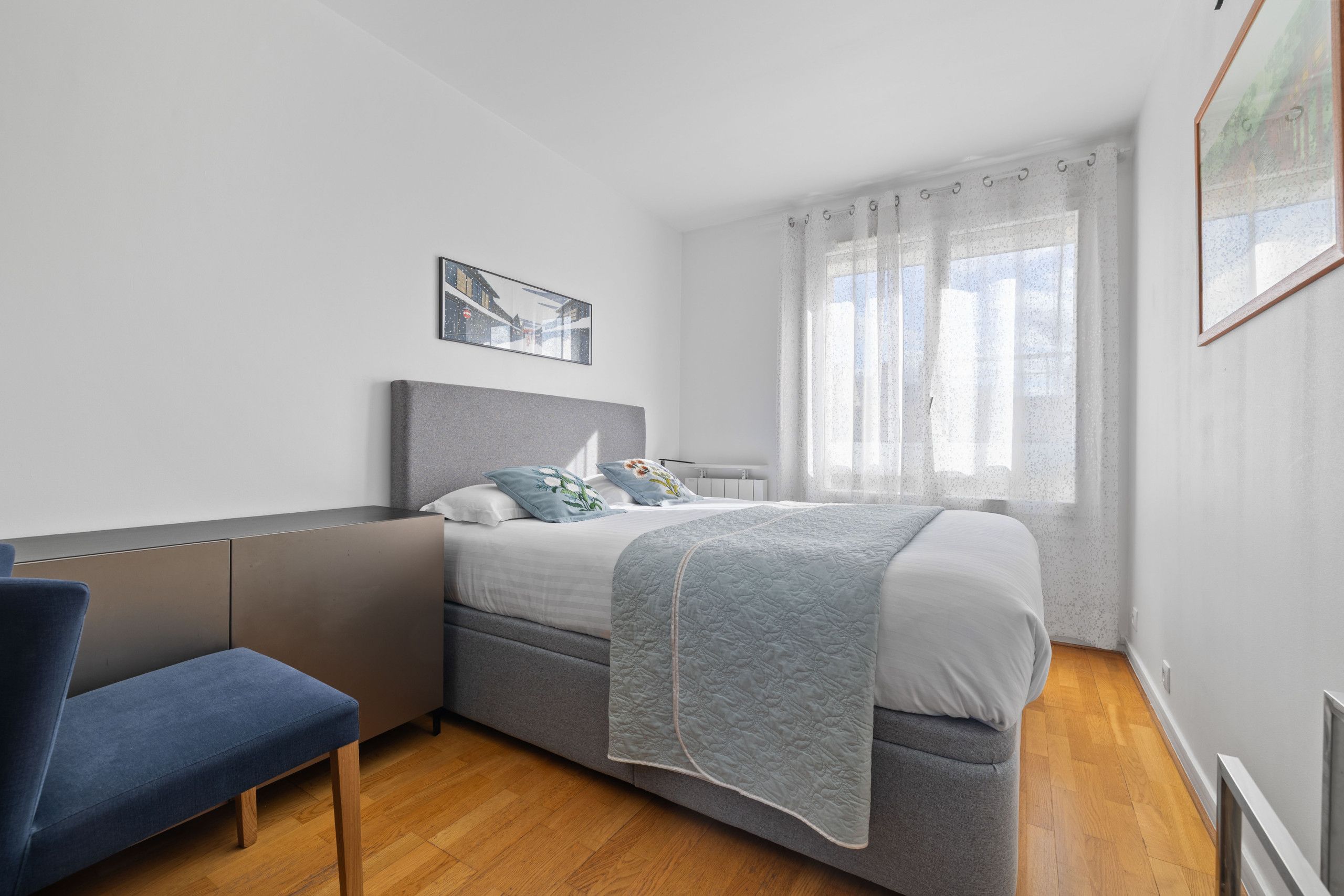 A modern bedroom with a grey bed, a blue chair, and a window with sheer curtains.