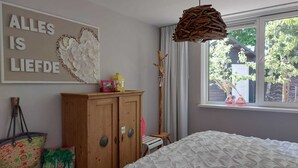 A bedroom with a bed, a wooden wardrobe, a window with a view of greenery, and a wall with a decorative piece and a sign. - Bungalow "The Shrimp" close to the beach! Air conditioning! South corner 1 (Renesse)