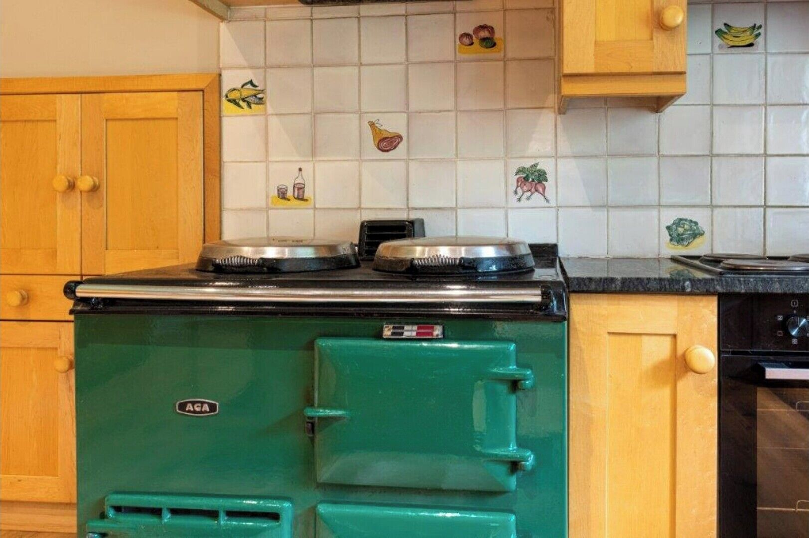 Une cuisine avec un poêle vert, des armoires en bois et un dosseret carrelé orné d’illustrations de produits alimentaires.