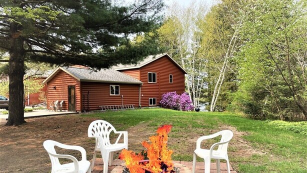 A wooden cabin with a brick fire pit in the center, surrounded by white plastic chairs. - Red Pines -  Elk Lake Condo #3 (Phillips)