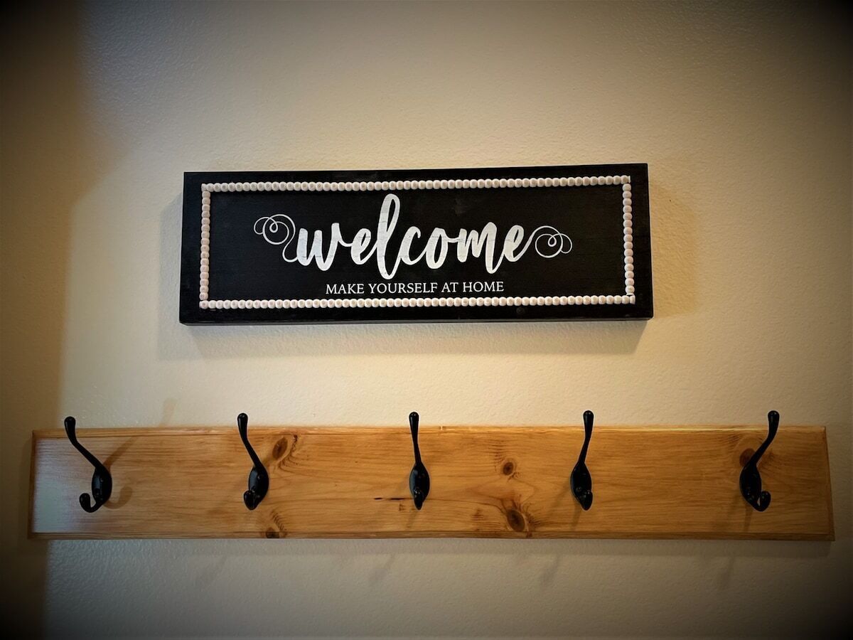Une pancarte d’accueil avec l’inscription « bienvenue, faites comme chez vous » est placée au-dessus d’un porte-manteau en bois muni de cinq crochets.