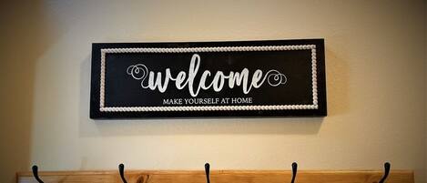 A welcome sign with the text "welcome make yourself at home" above a wooden coat rack with five hooks.