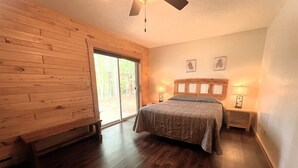 Ein Schlafzimmer mit Holzboden, einem Bett mit Kopfteil, zwei Nachttischen mit Lampen und einem Fenster mit Blick auf Bäume.