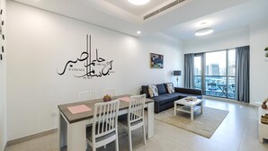 A modern living room with a dining area, a sofa, and a large window with a city view. - The Bay Residences - Canal View (Dubai)